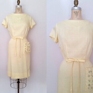 May include: A pale yellow vintage dress with a scoop neck, short sleeves, and a tie belt. The dress has a large, embellished pocket on the right side.