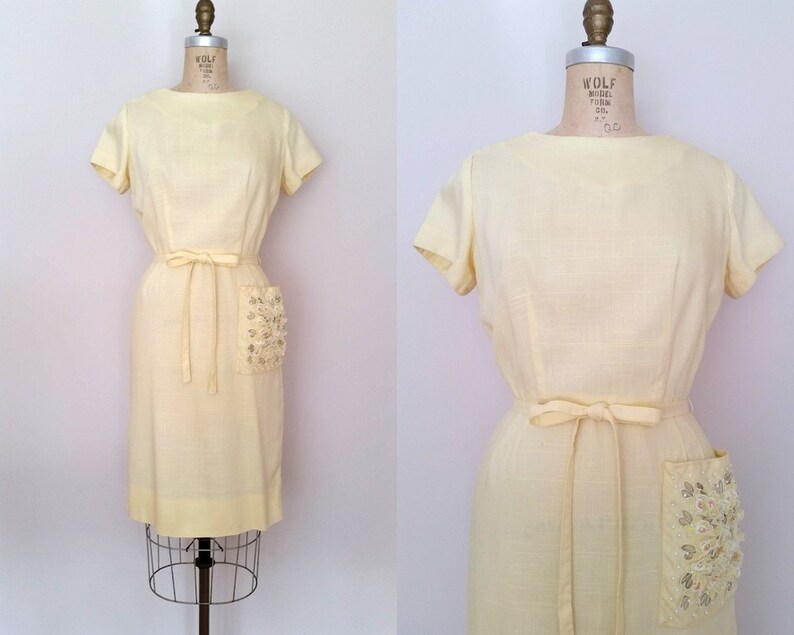 May include: A pale yellow vintage dress with a scoop neck, short sleeves, and a tie belt. The dress has a large, embellished pocket on the right side.