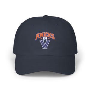 May include: A navy blue baseball cap featuring an embroidered design on the front. The text KNICKS IN is stitched in orange with a white outline, positioned above a large white letter V with a navy blue interior and a white border outline.