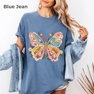 May include: A blue jean colored t-shirt featuring a large, central butterfly graphic. The butterfly is composed of various patterned patches in shades of pink, yellow, and light blue, accented with small white flowers and green leaves.