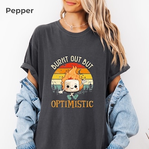 May include: Dark gray t-shirt with the text "BURNT OUT BUT OPTIMISTIC" and a graphic of a marshmallow on fire. The graphic has a sunset background with trees. The shirt is a casual, oversized fit.