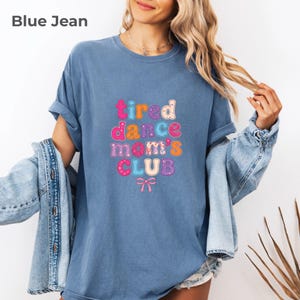 May include: A blue jean colored short-sleeved t-shirt featuring the text "tired dance mom's CLUB" in multi-colored, patterned bubble letters. Below the text is a small pink bow graphic. The shirt has a crew neckline and a relaxed fit.