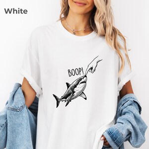 May include: A white t-shirt featuring a black and white graphic illustration of a shark being touched on the nose by a human finger. The text BOOP! is printed in black above the shark. The shirt has short sleeves and a crew neckline.