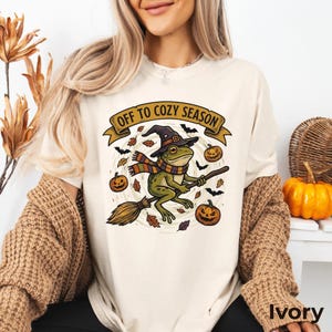 May include: An ivory t-shirt featuring a graphic of a green frog wearing a witch hat and a striped scarf while riding a broomstick. The design includes pumpkins, bats, and falling leaves with the text "OFF TO COZY SEASON" on a banner.