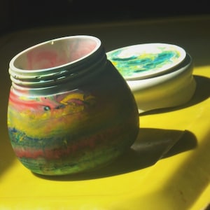 May include: A colorful, hand-painted jar with a matching lid. The jar features a swirl of red, yellow, green, and blue paint. The lid is white with a similar swirl of green and blue. The jar and lid sit on a yellow surface.