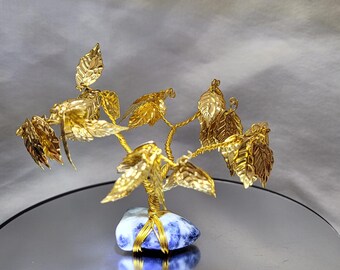 May include: A handcrafted golden wire tree sculpture with detailed leaves. The tree is mounted on a blue and white stone base, with the wire wrapped around it. The sculpture is displayed on a reflective surface.