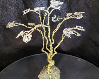 May include: A handcrafted wire tree sculpture with a gold-colored trunk and branches, and silver-colored leaves. The tree is rooted in a small, textured, gray stone. The sculpture is displayed on a reflective surface against a dark background.
