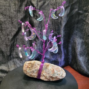 May include: A handcrafted wire tree sculpture with a purple trunk and branches. The branches hold clear, crescent-shaped ornaments with iridescent accents. The tree is secured to a stone base, creating a unique decorative piece.