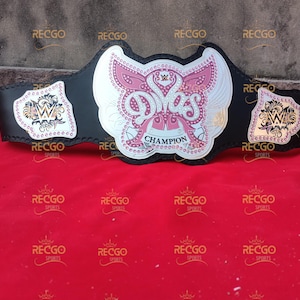Pode incluir: Um cinturão de campeonato de luta livre preto e prateado com um emblema central rosa e branco que diz "Divas Champion". O cinturão tem detalhes dourados e o logotipo da WWE nas placas laterais.