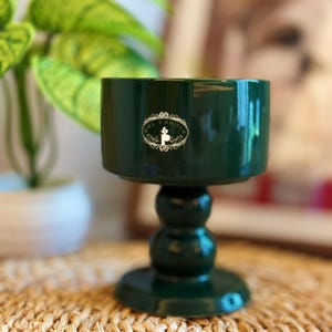 May include: A dark green candle holder with a round base and a pedestal design. The holder has a cylindrical top with a white and gold label that reads "OL CANDLE". The candle holder is sitting on a woven mat.