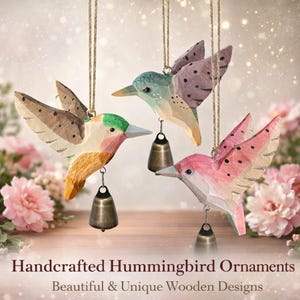May include: Three handcrafted wooden hummingbird ornaments in shades of green, pink, and brown, each with a small bell hanging below. The birds are suspended by twine. The text at the bottom reads "Handcrafted Hummingbird Ornaments".