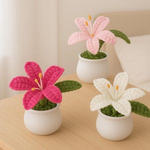 May include: Three crocheted lily flowers in small white pots. One is hot pink, one is light pink, and one is white. Each flower has green leaves and yellow stamens. The flowers sit on a light brown wooden surface.