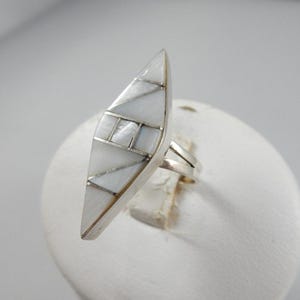 May include: A silver ring with a large, elongated, geometric design. The ring features a mosaic of white and silver elements, creating a striking visual contrast. The ring's design is composed of various shapes, including triangles and rectangles.