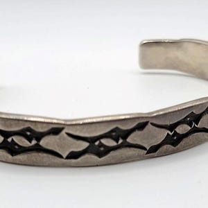 May include: Silver cuff bracelet with a repeating geometric pattern. The open-ended bracelet has a textured surface and a dark, etched design. The bracelet is displayed on a white background.