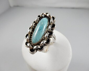 May include: A silver ring featuring an oval, light blue gemstone. The stone is set in a decorative, linked border. The ring is displayed on a white surface, showcasing its design and color.