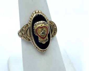 May include: Gold-toned ring with an oval black enamel center. The center features a red heart and a crest with the word "REGARD". The ring is displayed on a white stand.