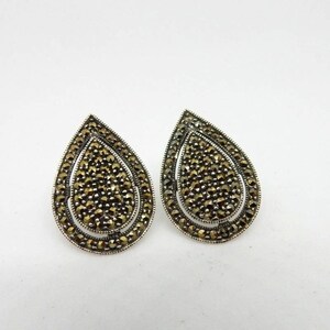 May include: Pair of teardrop-shaped stud earrings. Each earring features a silver-tone frame with a detailed interior filled with small, dark, sparkling stones. The earrings have a layered design, with the stones creating a textured surface.