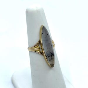 May include: A gold ring featuring a long, oval-shaped stone with gray and black inclusions. The ring's band has a decorative design with a split shank. The stone is set in a gold bezel.