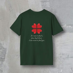 Puede incluir: Camiseta verde oscuro con un diseño de trébol de cuatro hojas rojo. El texto en la camiseta dice: "Es raro encontrar un trébol de cuatro hojas. Aún más raro encontrarte a ti."
