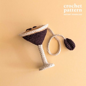 Pode incluir: Um copo de martini de crochê feito à mão com um corpo marrom escuro, uma borda bege e uma haste e base bege claro. O copo é conectado a uma azeitona marrom escura combinando por meio de um cordão bege. O texto "crochet pattern" e "instant download" são visíveis.