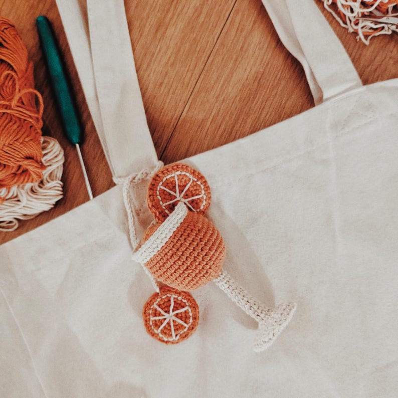 Op de afbeelding: Een witte draagtas met een handgemaakte oranje gehaakte cocktailbedel. De bedel heeft een gehaakt cocktailglas en twee sinaasappelschijfjes. Haakkatoen en een haaknaald zijn zichtbaar op de achtergrond.