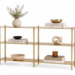 May include: A three-tier gold metal console table with thin vertical supports and flat shelves. The shelves hold decorative items including a beige vase with greenery, stacked books titled Kinfolk and AD at 100, bowls, a knot sculpture, and a candle.
