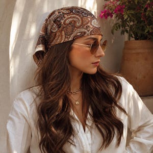 Puede incluir: Un pañuelo para la cabeza con un estampado de cachemira en tonos marrón, crema y negro, atado sobre el cabello largo y oscuro. Se combina con gafas de sol redondas de montura dorada con cristales tintados y un collar de cadena dorada.