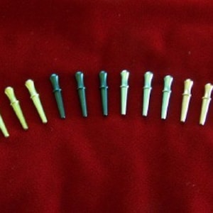 May include: A collection of twelve small, decorative pins. The pins are arranged in a row and feature a two-tone design with a rounded top and a slender, tapered body. The colors include yellow, dark green, and silver.