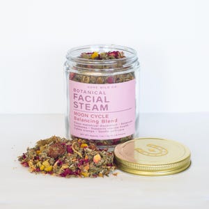 Könnte beinhalten: Ein Glas mit einer botanischen Mischung für Gesichtsdampfbäder, mit einem rosa Etikett mit der Aufschrift "Botanical Facial Steam Moon Cycle Balancing Blend". Das Glas ist offen, mit dem Deckel daneben. Ein kleiner Haufen der Mischung liegt neben dem Glas. Der Deckel ist goldfarben.
