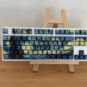 May include: A white mechanical keyboard with keycaps featuring a design inspired by Van Gogh's "Starry Night." The keycaps display a swirling pattern of blues, yellows, and blacks, creating a unique aesthetic. The keyboard is displayed on a small wooden easel.