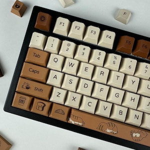 Puede incluir: Un teclado mecánico personalizado con marco negro y teclas color crema. Las teclas marrones presentan iconos de gatos y comida. La barra espaciadora tiene un diseño de gato marrón. Teclas adicionales están dispersas.