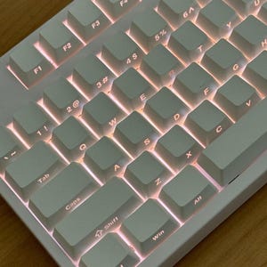 Puede incluir: Un teclado mecánico blanco con teclas iluminadas. Las teclas son de color gris claro con letras negras. El teclado tiene un diseño compacto, con teclas de función y una fila de números. Las teclas están retroiluminadas con un brillo cálido y anaranjado.