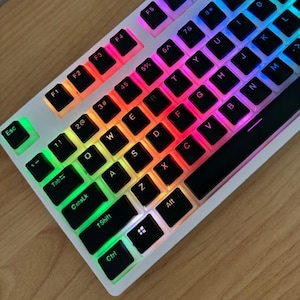 Può includere: Una tastiera meccanica bianca con tasti retroilluminati che mostrano un arcobaleno di colori. I tasti sono neri con lettere bianche. La tastiera è angolata su una superficie di legno.