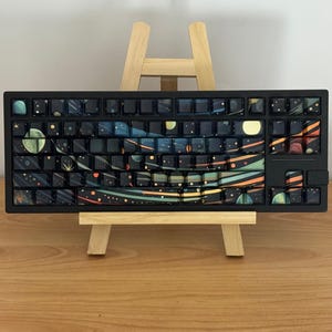 May include: Black mechanical keyboard with keycaps featuring a space-themed design. The keycaps display planets, stars, and nebulae in shades of blue, orange, and green. The keyboard is displayed on a small wooden easel.