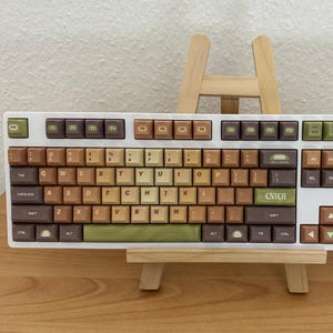 May include: A white mechanical keyboard with brown, green, and tan keycaps. The keyboard is displayed on a small wooden easel. The keycaps have a retro aesthetic, with a mix of colours and fonts.
