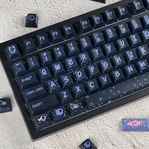 May include: A black mechanical keyboard with space-themed keycaps. The keycaps feature a dark blue background with white and light blue celestial designs, including planets, stars, and rockets. The keyboard has a standard layout with letters, numbers, and function keys.