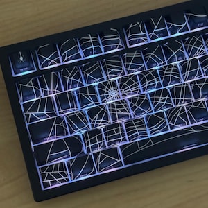 Puede incluir: Teclado mecánico negro con teclas de diseño de telaraña blanca. Las teclas están retroiluminadas con un arcoíris de colores. El teclado tiene una tecla WIN con un icono de araña.