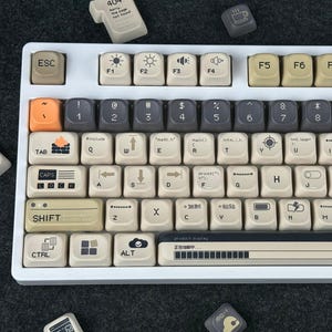 Könnte beinhalten: Eine weiße mechanische Tastatur mit beigefarbenen Tastenkappen und verschiedenen Symbolen und Texten. Die Tastatur hat eine Retro-Ästhetik mit einer Mischung aus grauen, orangefarbenen und schwarzen Tasten. Einige Tastenkappen haben Symbole, und die Tastatur enthält eine Anzeigeleiste.