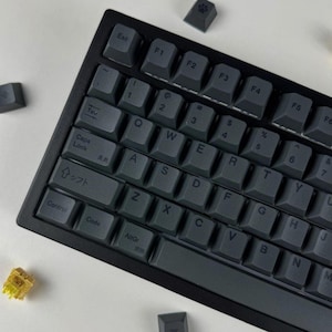 Könnte beinhalten: Eine schwarze mechanische Tastatur mit dunkelgrauen Tastenkappen. Mehrere Tastenkappen sind abgenommen, darunter eine mit einem Pfotenabdruck und eine mit einem Symbol. Die Tastatur hat ein Standard-QWERTZ-Layout mit japanischen Zeichen.