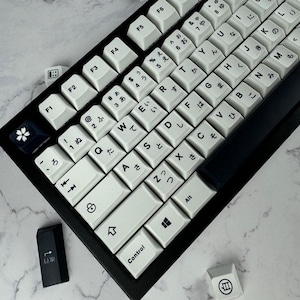 Może przedstawiać: Czarna klawiatura mechaniczna z białymi nakładkami klawiszy z japońskimi znakami i angielskimi literami. Klawiatura ma czarną ramę i kilka luźnych nakładek klawiszy. Nakładki klawiszy mają czysty, minimalistyczny design.