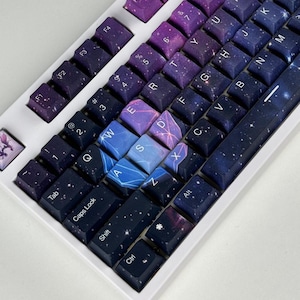 May include: A white mechanical keyboard with custom keycaps. The keycaps feature a galaxy-themed design with shades of purple, blue, and white. The keyboard layout includes standard keys, function keys, and a space bar.