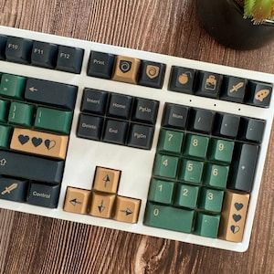 Może przedstawiać: Biała klawiatura mechaniczna z niestandardowymi nakładkami na klawisze. Nakładki na klawisze są mieszanką ciemnozielonego, czarnego i złotego. Niektóre klawisze mają ikony, takie jak serca, bomby i miecze. Klawiatura leży na drewnianej powierzchni.