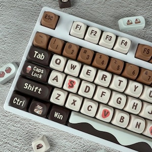 May include: A white mechanical keyboard with custom keycaps. The keycaps are a mix of white and chocolate brown, with some keys featuring dessert-themed icons. The keyboard has a unique design, with the letters and symbols in a playful font.