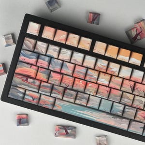 Könnte beinhalten: Eine schwarze mechanische Tastatur mit individuellen Tastenkappen, die eine Landschaft in Orange, Rosa, Blau und Grau zeigen. Mehrere lose Tastenkappen mit dem gleichen Design sind um die Tastatur herum verstreut.