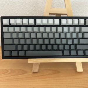 May include: A mechanical keyboard with a black frame and a gradient keycap design. The keycaps transition from white to gray to black. The keyboard is displayed on a small wooden easel, set against a neutral background.