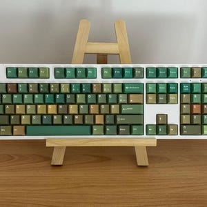 May include: A white mechanical keyboard with green and brown keycaps. The keyboard is displayed on a small wooden easel. The keycaps have a variety of green and brown shades, creating a unique aesthetic. The keyboard has a number pad.