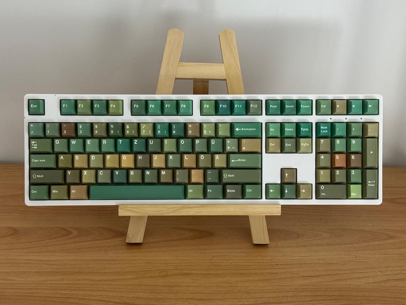 May include: A white mechanical keyboard with green and brown keycaps. The keyboard is displayed on a small wooden easel. The keycaps have a variety of green and brown shades, creating a unique aesthetic. The keyboard has a number pad.