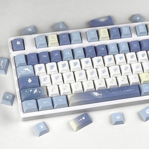 Può includere: Una tastiera meccanica bianca con tasti personalizzati in tonalità blu e bianco. I tasti presentano disegni a tema acquatico, tra cui pesci, onde e stelle. La barra spaziatrice ha la scritta "Aquarium of dreams".