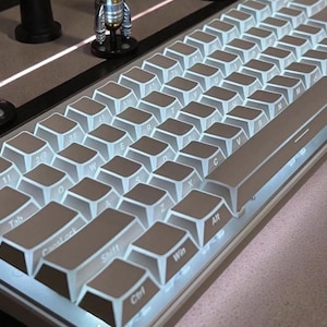 May include: A close-up of a mechanical keyboard with white keycaps and a gray frame. The keys are backlit with a soft white glow. The keyboard has a compact layout, with keys for Tab, CapsLock, Shift, Ctrl, Win, and Alt.