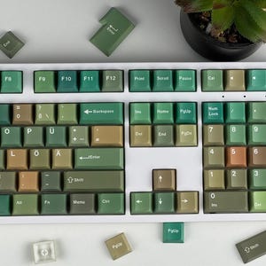 May include: A full-sized mechanical keyboard with a white frame and green, tan, and brown keycaps. The keycaps are arranged in a standard QWERTY layout, with function keys and a number pad. Several loose keycaps are scattered around the keyboard.